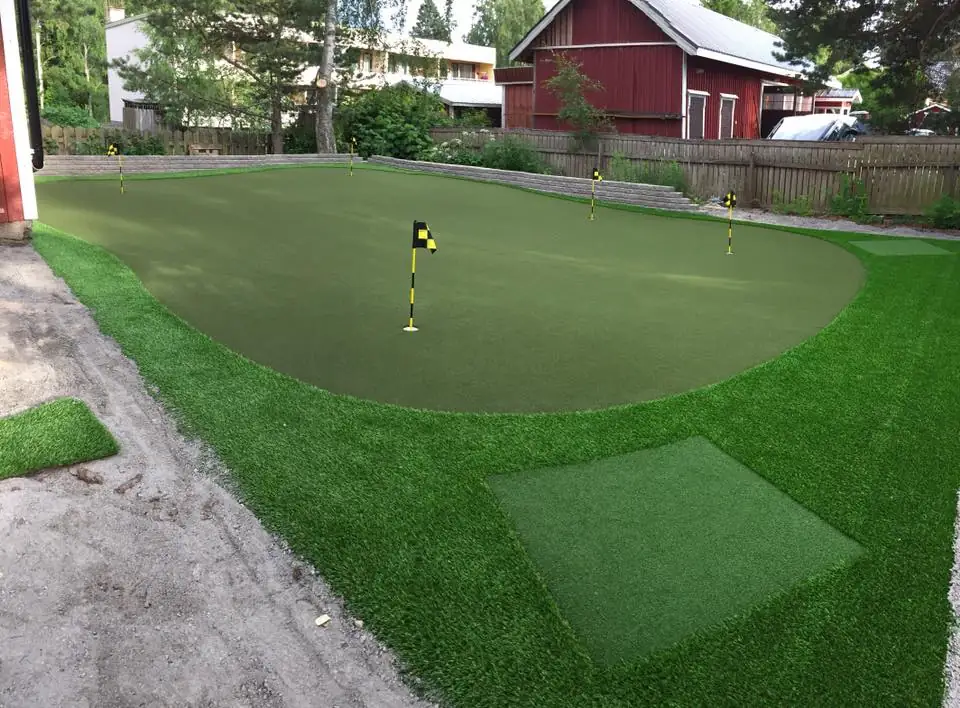Viheriö kahdella lipulla ja reunanurmeen upotettu lyöntimatto, taustalla punainen aitta.