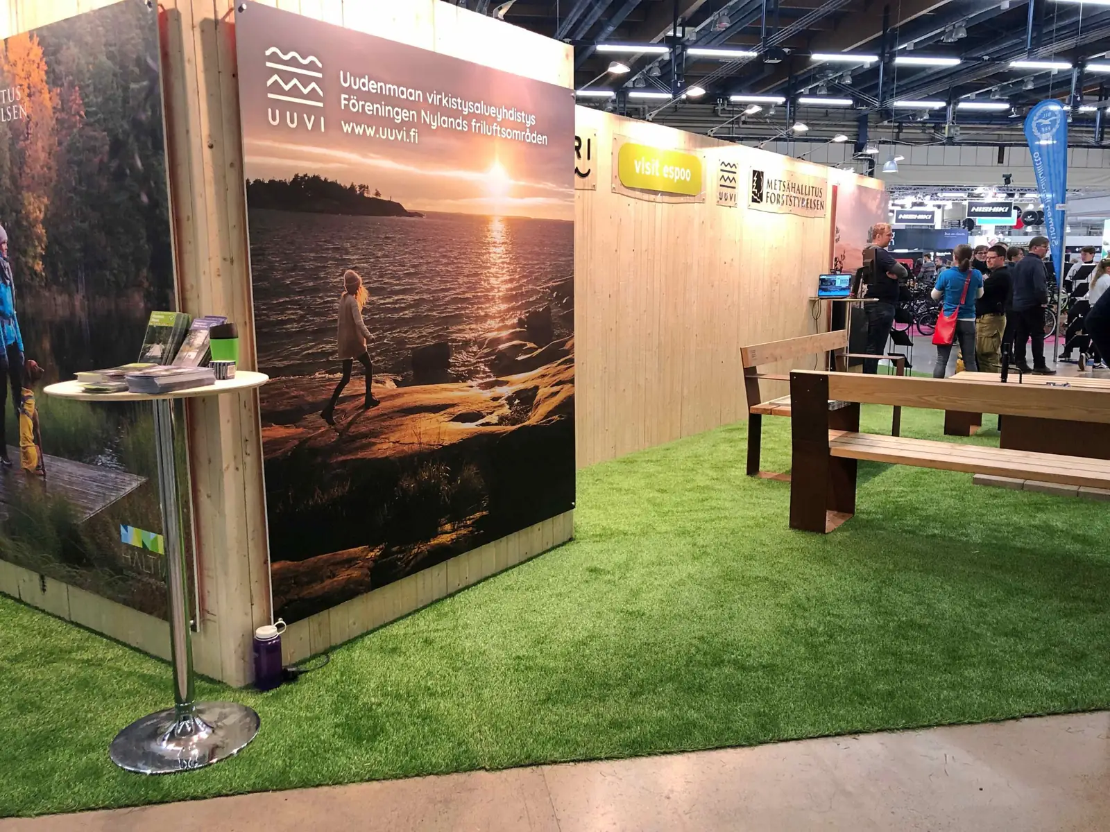 Messuosasto, jonka lattiana on vihreä tekonurmi. Osastolla pöytä, penkit ja esitetelineet.