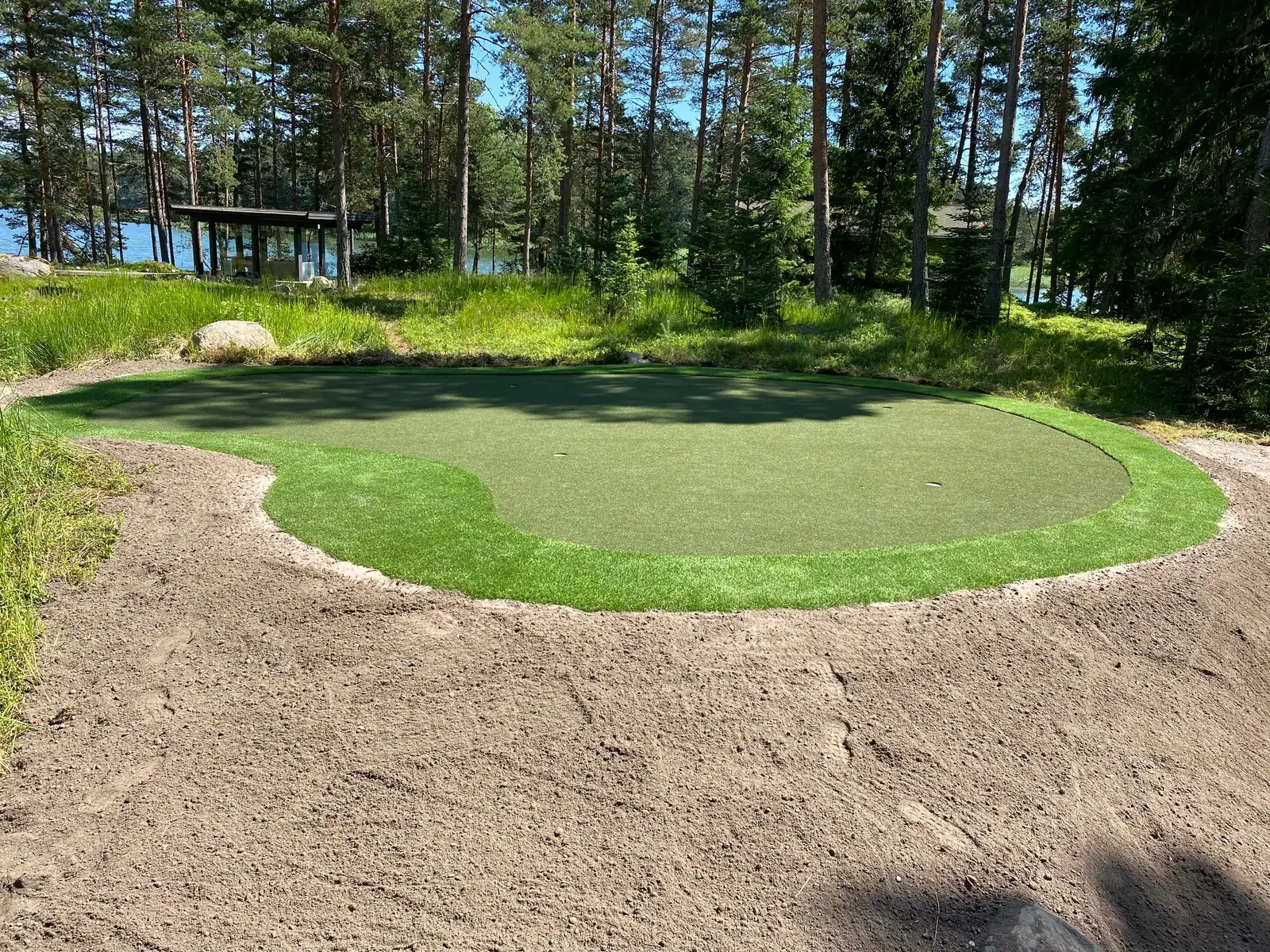 Tekonurmiviheriö ja sen ympärille muotoiltu hiekka-alue mäntymetsässä järven rannalla.