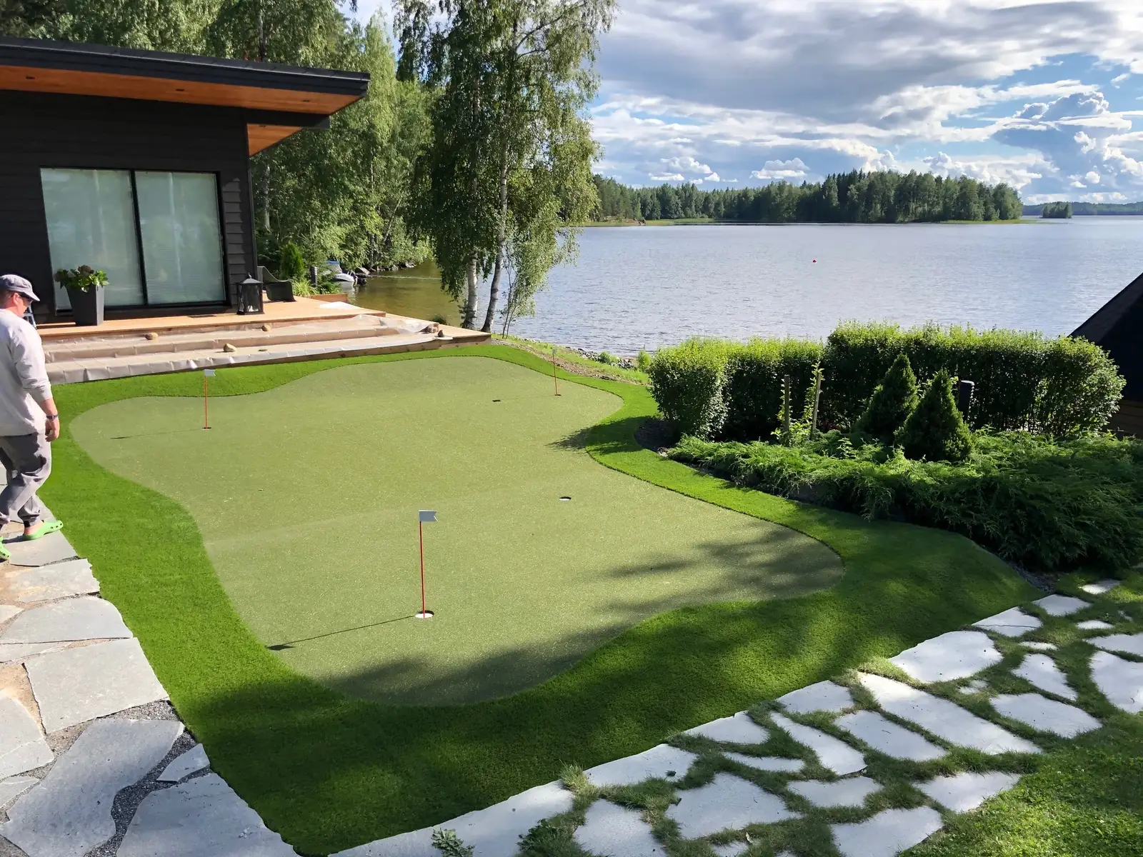 Golfviheriö järven rannalla mökin edessä: puttiviheriö, lippu ja kaareva reunanurmi, reunassa liuskekivipolku.